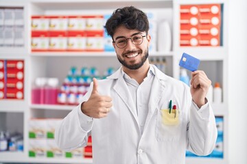 Canvas Print - Hispanic man with beard working at pharmacy drugstore holding credit card smiling happy and positive, thumb up doing excellent and approval sign