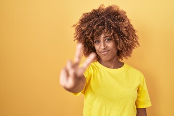 Sticker - Young hispanic woman with curly hair standing over yellow background smiling looking to the camera showing fingers doing victory sign. number two.