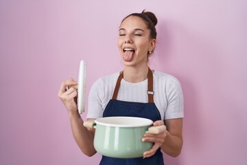 Poster - Young hispanic girl wearing apron holding cooking pot sticking tongue out happy with funny expression.