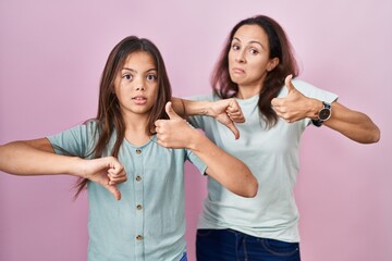 Wall Mural - Young mother and daughter standing over pink background doing thumbs up and down, disagreement and agreement expression. crazy conflict