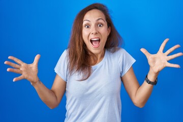 Poster - Brunette woman standing over blue background celebrating crazy and amazed for success with arms raised and open eyes screaming excited. winner concept