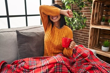 Canvas Print - Young hispanic woman sitting on the sofa drinking a coffee at home smiling cheerful playing peek a boo with hands showing face. surprised and exited