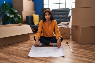 Young hispanic woman sitting on the floor at new home looking at blueprints thinking attitude and sober expression looking self confident