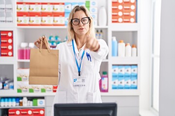 Sticker - Young blonde woman working at pharmacy drugstore holding paper bag pointing with finger to the camera and to you, confident gesture looking serious