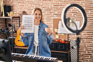 Sticker - Young caucasian woman doing online music tutorial showing music sheet smiling happy and positive, thumb up doing excellent and approval sign