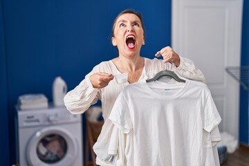 Poster - Middle age hispanic woman holding shirt on hanger and detergent powder angry and mad screaming frustrated and furious, shouting with anger looking up.