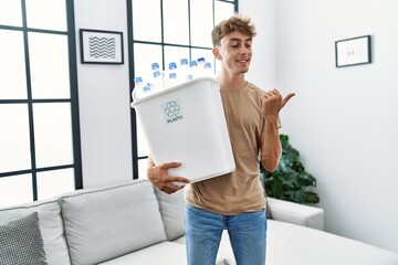 Poster - Young caucasian man holding wastebasket with recycling plastic bottles at home pointing thumb up to the side smiling happy with open mouth