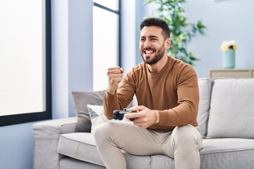 Poster - Young hispanic man playing video game sitting on sofa at home