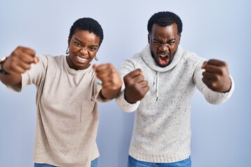 Canvas Print - Young african american couple standing over blue background together angry and mad raising fists frustrated and furious while shouting with anger. rage and aggressive concept.