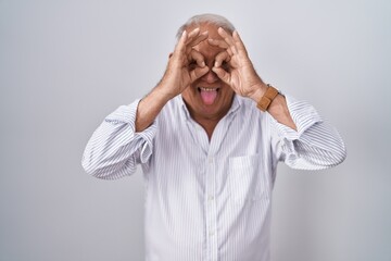 Sticker - Senior man with grey hair standing over isolated background doing ok gesture like binoculars sticking tongue out, eyes looking through fingers. crazy expression.