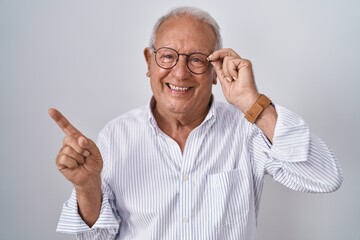 Poster - Senior man with grey hair holding glasses with hand smiling happy pointing with hand and finger to the side