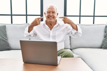 Wall Mural - Senior man using laptop at home sitting on the sofa smiling cheerful showing and pointing with fingers teeth and mouth. dental health concept.