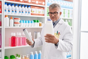 Poster - Middle age grey-haired man pharmacist holding shampoo bottles at laboratory