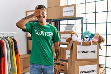 Sticker - Young handsome hispanic man wearing volunteer t shirt at donations stand smiling and laughing with hand on face covering eyes for surprise. blind concept.