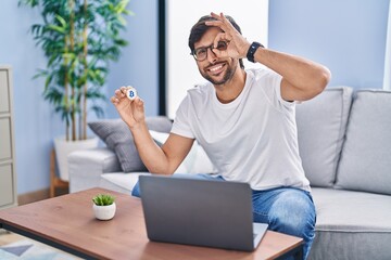 Sticker - Handsome latin man holding virtual currency bitcoin using laptop smiling happy doing ok sign with hand on eye looking through fingers