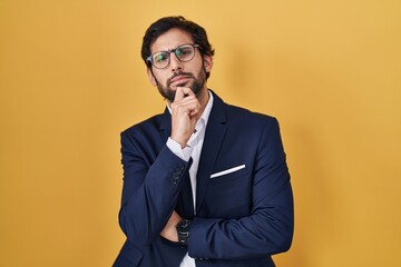 Canvas Print - Handsome latin man standing over yellow background looking confident at the camera smiling with crossed arms and hand raised on chin. thinking positive.