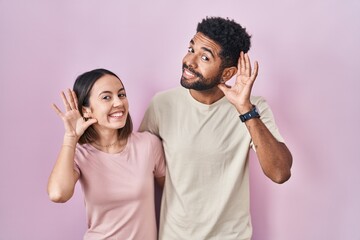 Poster - Young hispanic couple together over pink background smiling with hand over ear listening an hearing to rumor or gossip. deafness concept.