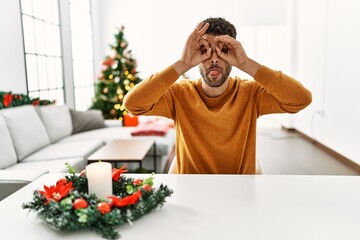 Sticker - Arab young man sitting on the table by christmas tree doing ok gesture like binoculars sticking tongue out, eyes looking through fingers. crazy expression.