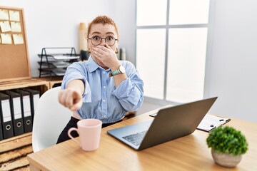 Wall Mural - Young redhead woman working at the office using computer laptop laughing at you, pointing finger to the camera with hand over mouth, shame expression