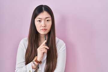 Canvas Print - Chinese young woman holding pencil over pink background relaxed with serious expression on face. simple and natural looking at the camera.