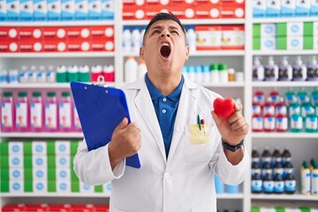 Sticker - Young hispanic man working at pharmacy drugstore holding heart angry and mad screaming frustrated and furious, shouting with anger looking up.