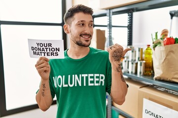 Canvas Print - Young volunteer man holding your donation matters banner pointing thumb up to the side smiling happy with open mouth