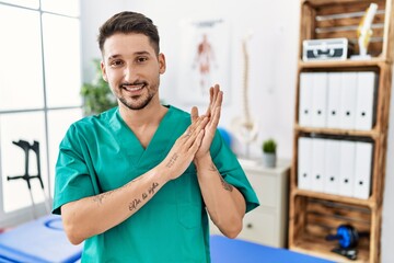 Canvas Print - Young physiotherapist man working at pain recovery clinic clapping and applauding happy and joyful, smiling proud hands together