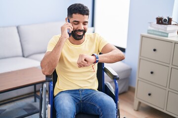 Sticker - Young arab man talking on smartphone looking watch sitting on wheelchair at home