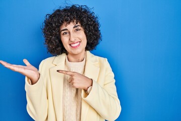 Sticker - Young brunette woman with curly hair standing over blue background amazed and smiling to the camera while presenting with hand and pointing with finger.