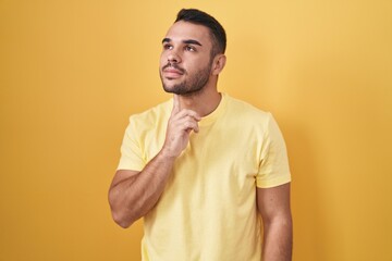 Wall Mural - Young hispanic man standing over yellow background thinking concentrated about doubt with finger on chin and looking up wondering