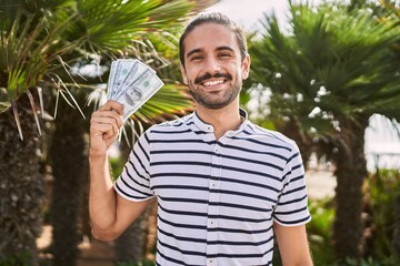 Wall Mural - Young hispanic man holding dollars close to face looking positive and happy standing and smiling with a confident smile showing teeth