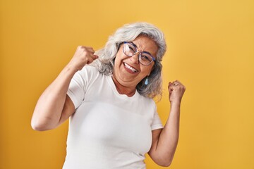 middle age woman with grey hair standing over yellow background very happy and excited doing winner 