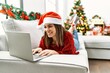 © Krakenimages.com - Young hispanic girl using laptop lying on the sofa by christmas tree at home.