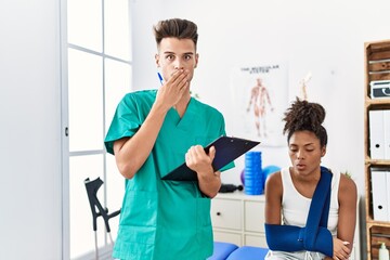 Canvas Print - Physiotherapist working with patient wearing arm on sling at the clinic covering mouth with hand, shocked and afraid for mistake. surprised expression