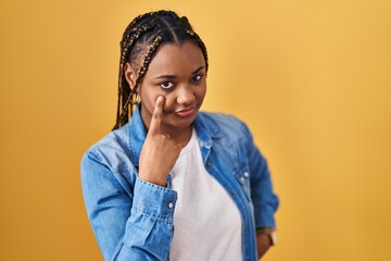 Sticker - African american woman with braids standing over yellow background pointing to the eye watching you gesture, suspicious expression