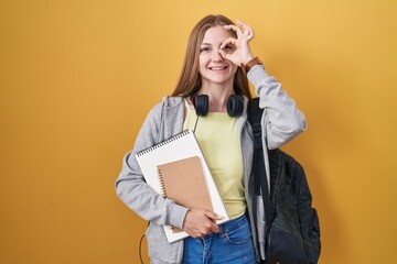 Canvas Print - Young caucasian woman wearing student backpack and holding books doing ok gesture with hand smiling, eye looking through fingers with happy face.