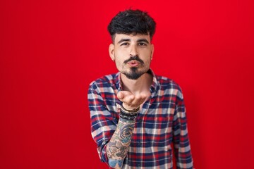 Poster - Young hispanic man with beard standing over red background looking at the camera blowing a kiss with hand on air being lovely and sexy. love expression.