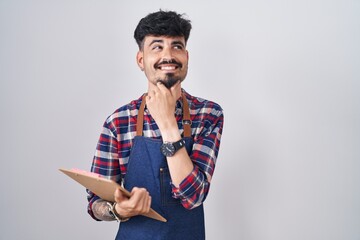 Wall Mural - Young hispanic man with beard wearing waiter apron holding clipboard with hand on chin thinking about question, pensive expression. smiling and thoughtful face. doubt concept.