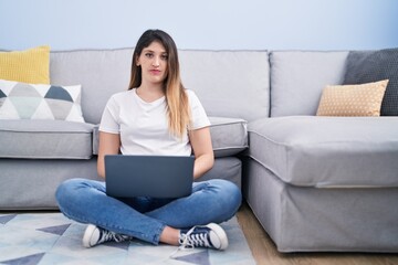 Sticker - Young brunette woman sitting on the floor at home using laptop thinking attitude and sober expression looking self confident