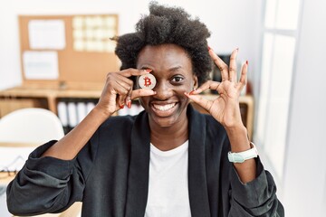 Wall Mural - African young woman holding bitcoin on eye at the office doing ok sign with fingers, smiling friendly gesturing excellent symbol