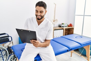 Sticker - Young arab man wearing physiotherapist uniform writing on clipboard at clinic