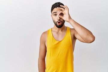 Canvas Print - Young handsome man with beard standing over isolated background doing ok gesture shocked with surprised face, eye looking through fingers. unbelieving expression.