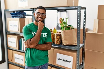 Poster - Young indian man volunteer holding donations box looking confident at the camera smiling with crossed arms and hand raised on chin. thinking positive.