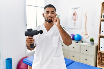 Poster - Young indian physiotherapist holding therapy massage gun at wellness center mouth and lips shut as zip with fingers. secret and silent, taboo talking