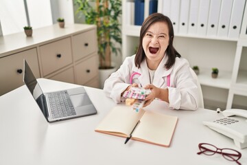 Poster - Hispanic girl with down syndrome wearing doctor uniform and stethoscope holding pills angry and mad screaming frustrated and furious, shouting with anger. rage and aggressive concept.