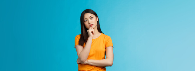 Wall Mural - Thoughtful dreamy smart asian girl thinking, making plan inside head, touch chin, look up focused, contemplating interesting scene, pondering, making choice, taking decision, stand blue background