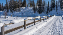 An Old Bridge Over A Frozen River. Tire Tracks Are Trampled In The Snow. There Is A Layer Of Snow On The Unpainted Wooden Railing. Coniferous Trees On The Hill. Clear Blue Sky. Altai