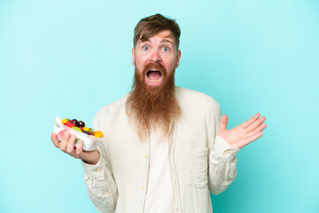 Wall Mural - Redhead man with long beard holding a bowl of fruit isolated on blue background with shocked facial expression