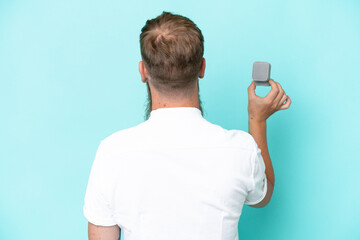 Wall Mural - Redhead man with long beard holding a engagement ring isolated on blue background in back position