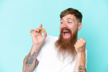 Wall Mural - Redhead man with long beard holding a Bitcoin isolated on blue background celebrating a victory